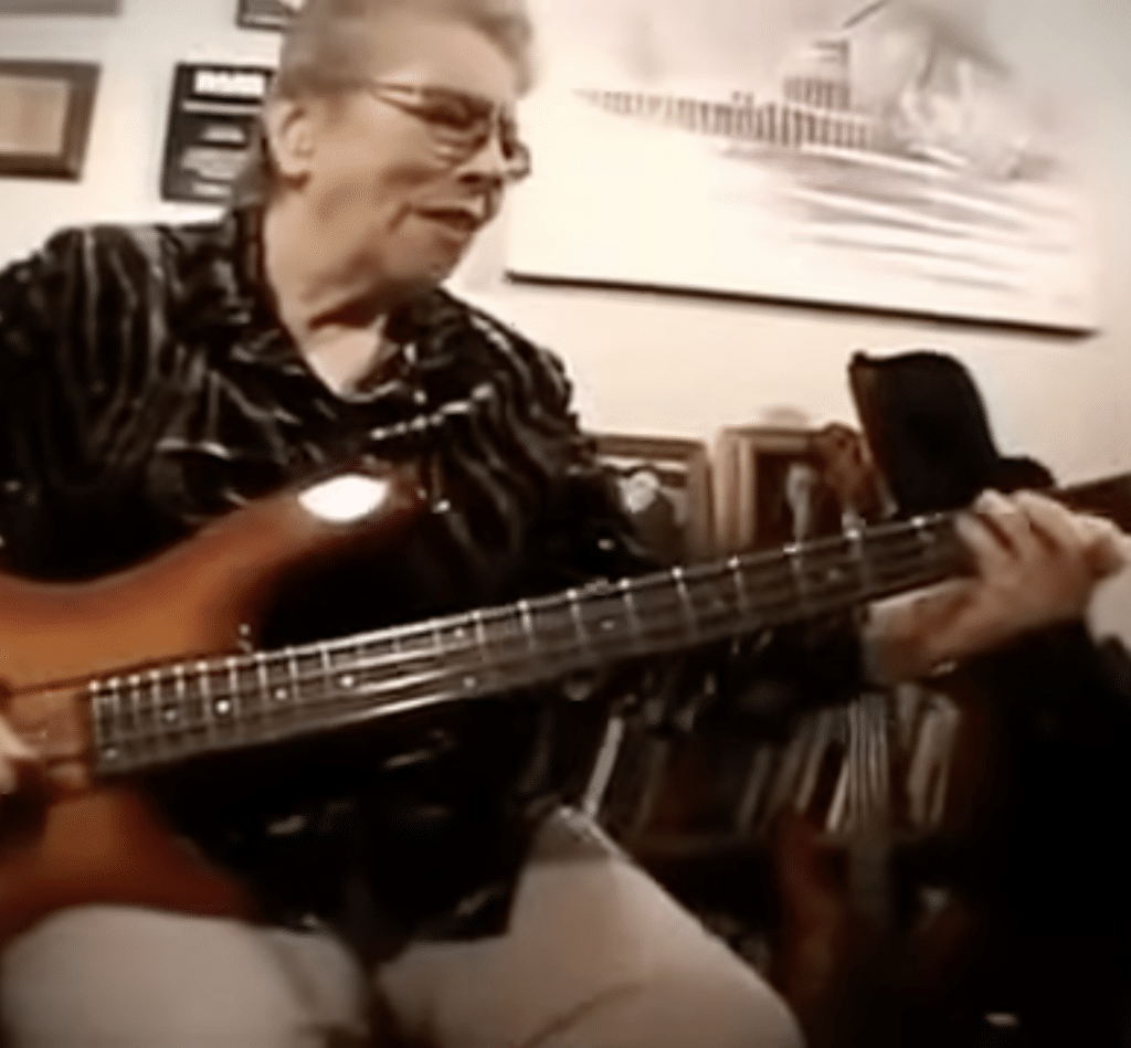 Carol Kaye plays a guitar while sitting on a couch in a picture-framed room.